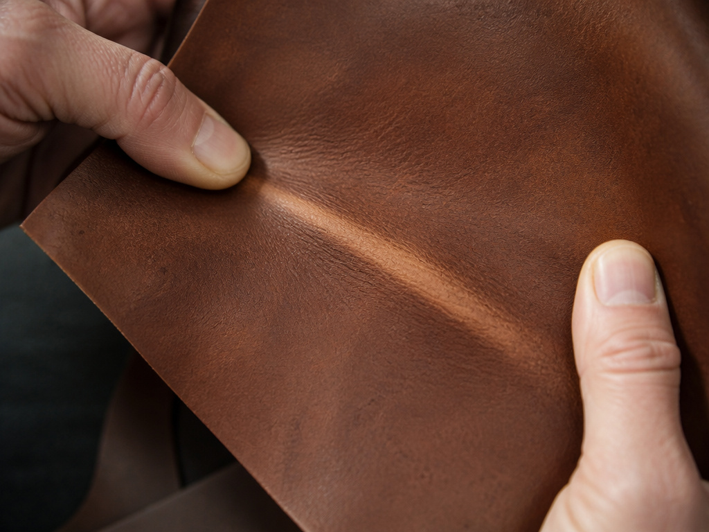 Close-up hands testing semi-aniline leather's pull-up effect and natural grain.