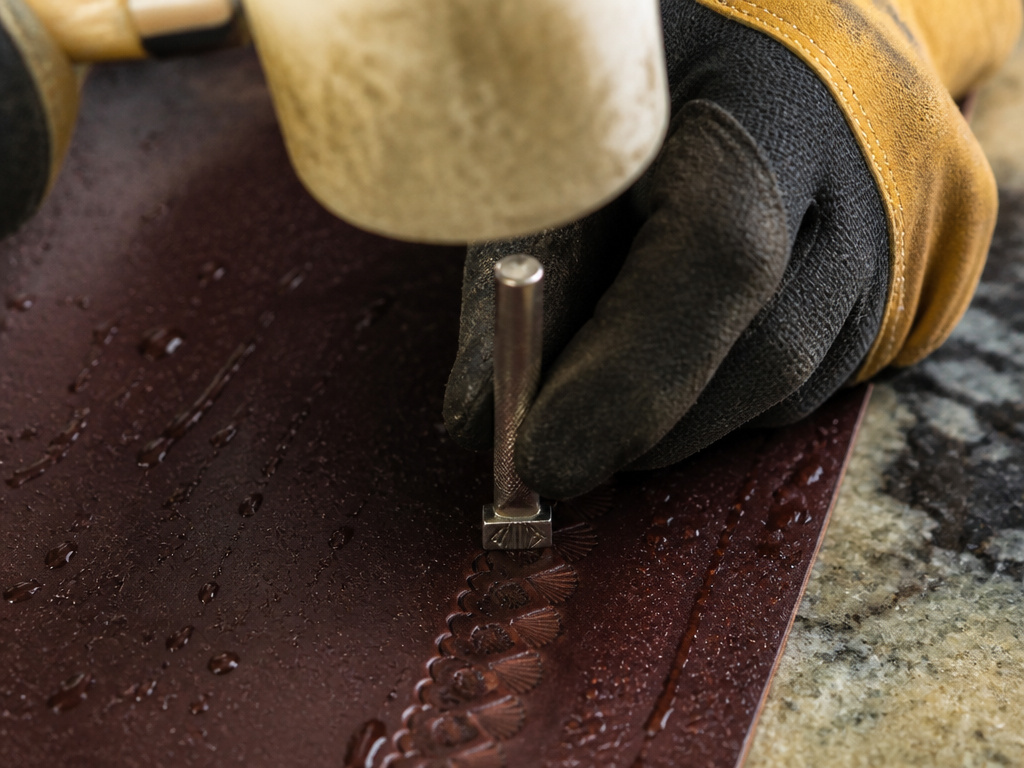 Close-up of hands using a mallet to strike a metal stamp for leather embossing.